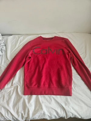 Röd sweatshirt från Calvin Klein - Röd sweatshirt från Calvin Klein med stort tryck 'Calvin' på bröstet. Klassisk rund halsringning och långa ärmar. Tillverkad i mjukt material som passar perfekt till en avslappnad stil.