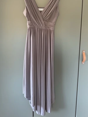 Grå långklänning Chiara Forthi - Elegant grå långklänning från Chiara Forthi med omlott upptill och vackert fallande kjol. Klänningen har breda axelband och markerad midja som ger en smickrande siluett. Perfekt för dig som vill ha en stilren och klassisk look.