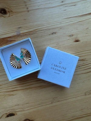 Guldörhängen Caroline Svedbom - Säljer ett par öronhänge från Caroline Svedbom i guld med solfjädersform och gröna/turkosa kristaller. Helt nya aldrig använda💕