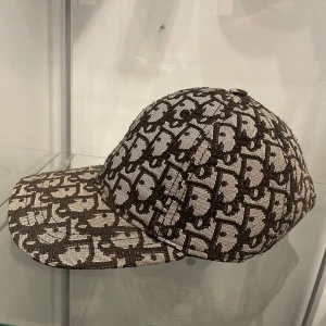 Mönstrad keps från Christian Dior - Snygg keps från Christian Dior med ett grått och beige mönster över hela kepsen. Justerbar rem baktill för perfekt passform. Tillverkad i 100% bomull och har en klassisk böjd skärm.