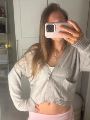 Grå cropped kofta med knappar - Mysig grå cropped kofta med tre knappar framtill och två stora fickor. Koftan har ribbade muddar och är tillverkad i ett mjukt, frottéliknande material. Perfekt för lager på lager och chill dagar.