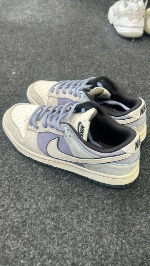 Nike Dunk Low i lila och grå mocka - Nike Dunk Low sneakers i en snygg mix av lila och ljusgrå mocka med svart swoosh och vita detaljer. Skorna har perforerad tåbox, klassiska snörningar och svart innerfoder. Perfekt för dig som gillar streetwear och vill sticka ut med stilrena färger.