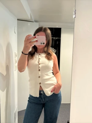 Vit ribbad ärmlös topp med knappar - Säljer en stilren vit ribbad topp med knappar framtill. Toppen är ärmlös och har en rak passform som ger en clean look. Perfekt att matcha med jeans eller kjol för en enkel men snygg outfit.