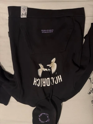 Svart hoodie från Hocrich med tryck - Säljer en svart hoodie från Hocrich med stor magficka och tryck av två vita fåglar samt texten 'HOCRICH' på framsidan. På huvan finns ett broderat motiv och text. Hoodien har dragsko och är gjord i mjukt bomullsmaterial.