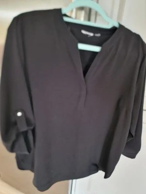 Svart blus med v-ringning - Stilren svart blus med v-ringning och trekvartsärmar som kan rullas upp och fästas med knapp. Lös passform och mjukt, lätt material som ger en avslappnad look. Perfekt att matcha med jeans eller kjol för en trendig outfit. Osäker storlek L eller XL. Lappen är borta. Vet ej märke