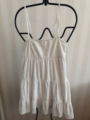 Vit somrig klänning med spetsdetaljer - Söt vit kortklänning med tunna axelband och volangkant. Klänningen har broderade spetsdetaljer och är tillverkad i ett lätt bomullstyg, perfekt för varma dagar. Passar dig som gillar en luftig och romantisk stil.