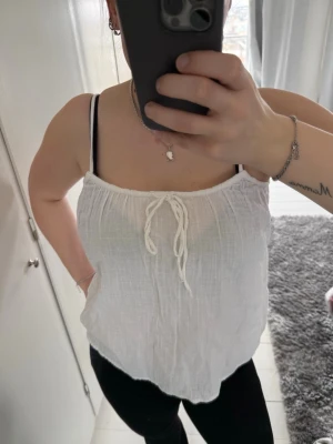 Vit linnetopp med knytband - Säljer en luftig vit linnetopp med tunna axelband och knytband framtill. Toppen har en avslappnad passform och är gjord i ett mjukt, strukturerat material som känns perfekt för varma dagar.