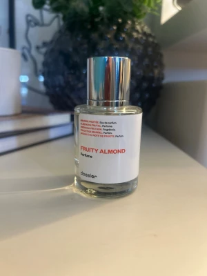 Fruity Almond parfym från Dossier - En stilren parfymflaska från Dossier med doften Fruity Almond. Flaskan är genomskinlig med en vit etikett och har ett modernt, silverfärgat lock. Designen är minimalistisk och passar perfekt på sminkbordet. Parfymen har en fräsch och fruktig mandeldoft. Endast testad