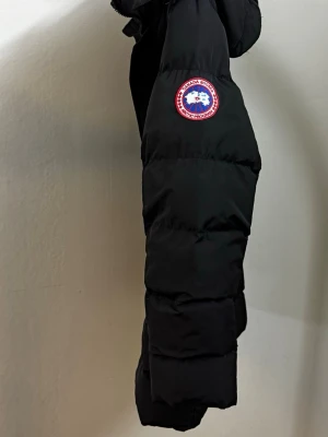 Svart dunjacka från Canada Goose - Säljer en svart dunjacka från Canada Goose med avtagbar huva och fuskpäls. Jackan har två stora dragkedjeförsedda fickor framtill och klassisk Canada Goose-logga på ärmen. Perfekt för kalla vinterdagar och riktigt varm.