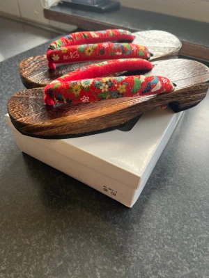 Japanska geta sandaler med blommigt band - Unika japanska geta sandaler i mörkt trä med traditionella röda tygband dekorerade med färgglada blommönster. Snyggt handgjorda och perfekta för dig som vill sticka ut med en klassisk och cool stil. Passar till både streetwear och mer traditionella outfits.