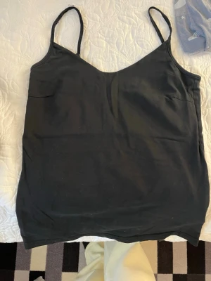 Svart linnetopp med smala axelband - Säljer en enkel svart linnetopp med smala axelband och v-ringad hals. Toppen har en rak passform och är ärmlös, perfekt att styla till olika outfits. Materialet känns mjukt och lätt, troligen bomull eller polyester.