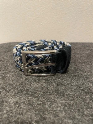 Flätat blått och grått bälte, storlek 100-105 cm - Snyggt flätat bälte i blått, grått och vitt med silverfärgat spänne. Bältet har en flexibel och justerbar design som gör det enkelt att anpassa till olika outfits. Avgör själv hur lång det är med hjälp av bilden. 