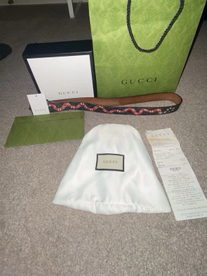 Gucci svart bälte med orm-mönster - Gucci bälte i svart skinn med ett coolt orm-mönster i rött och vitt. Bältet kommer med originalpåse, box och påse från Gucci. Perfekt accessoar för att lyfta din outfit och ge en lyxig touch. Detta bältet är otroligt snyggt, och passar perfekt till sommaren, Det är inte allt många skador förutom att färgen går upp lite  ( fler bilder i dm) jag har box påse,kvitto som tyder på att bältet är äkta priset är inte hugget i sten . Funderingar? Dm! Tack i förväg 