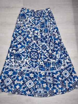 Blåvit långkjol med blommigt mönster - Säljer en långkjol i blått och vitt med ett detaljerat blommigt mönster. Kjolen har en lätt utsvängd form och är tillverkad i ett mjukt, tunt material som passar perfekt för varmare dagar. Ingen synlig dragkedja eller knappar. Perfekt för dig som gillar boho-stil!