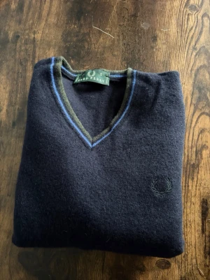 Fred Perry Tröja - Fred Perry stickad tröja i storlek S. Klassisk V-neck i mörk färg med subtil logga och fina detaljer vid kragen. Tillverkad i kashmir vilket ger en mjuk och premium känsla. Bra skick utan större defekter. Passformen är något mindre i storlek. Om du lyckas få den tillbaka i storlek så har du ett fynd. Perfekt för en clean casual/old money stil.