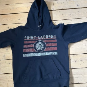 Mörkblå Saint Laurent hoodie - Säljer en mörkblå hoodie från Saint Laurent med tryck i vitt och rött på bröstet. Hoodien har huva med snören och en stor magficka. Perfekt för dig som gillar streetstyle och vill ha något snyggt och bekvämt.