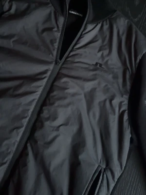 Svart hybrid jacka från J.Lindberg  - Snygg svart bomberjacka med dragkedja framtill och ribbade muddar. Jackan har två sidofickor och en extra ficka med dragkedja på ärmen. Materialet är glansigt och ger en klassisk streetwear-look.