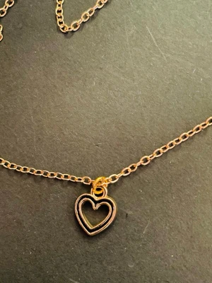 By prisförslag- Guldfärgat halsband 45 cm med hjärthänge - Snyggt halsband i guldfärg med en tunn kedja och ett litet hjärta som hänge. Hjärtat är öppet i mitten och har en minimalistisk design. Perfekt accessoar för att lyfta din outfit med en söt detalj. Materialet ser ut att vara metall. För längre kedja 1:-/cm extra. 