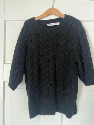 Svart stickad tröja med pärlor Zara - Snygg svart stickad tröja från Zara med pärldetaljer över hela tröjan. Tröjan har ett kabelstickat mönster, rund halsringning och trekvartsärmar. Ribbstickade muddar vid ärmslut och nederkant. Perfekt för dig som gillar detaljer och vill ha något extra.