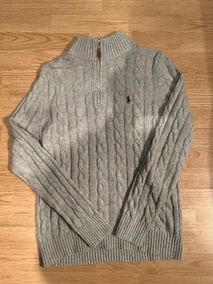 Grå stickad tröja Polo Ralph Lauren - Snygg grå kabelstickad tröja från Polo Ralph Lauren med halv dragkedja och ribbad krage. Tröjan har det klassiska broderade logotypen på bröstet och är tillverkad i mjukt material. Perfekt för dig som gillar stilrena och tidlösa plagg.