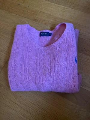 Rosa kabelstickad tröja Polo Ralph Lauren - Snygg rosa kabelstickad tröja från Polo Ralph Lauren med rund halsringning och blå broderad logga på bröstet. Tröjan har långa ärmar och ribbade muddar vid ärmslut och nederkant. Perfekt för dig som gillar klassisk preppy stil.