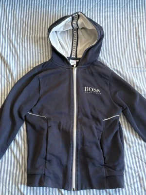 Mörkblå hoodie från Hugo Boss - Snygg mörkblå hoodie från Hugo Boss med vit dragkedja och logga på bröstet. Huvan har vit insida och detaljer med BOSS-tryck längs kanten. Praktiska fickor framtill och diskreta vita linjer vid fickorna. Perfekt för en stilren och avslappnad look. Storlek XS i barnstorlek.