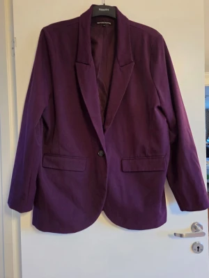 Lila kavaj, stl 44 - Stilren lila kavaj med klassisk krage, enkelknäppning och två fickor framtill. Kavajen har långa ärmar med knappdetaljer vid ärmslut och är tillverkad i ett mjukt tyg som ger en snygg passform.