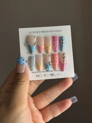 Färgglada press-on naglar med stenar - Säljer ett set press-on naglar i mandelform med cool design i blått, rosa och gult. Naglarna har prickiga detaljer, silverfärgade stenar och mixade färgblock. Materialet är plast och de är handgjorda för en unik look.