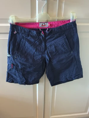 Mörkblå shorts från McKinley - Mörkblå shorts från McKinley med rosa innerfoder och detaljer. Shortsen har flera fickor, dragkedja på ena benet och bälteshällor. Tillverkade i ett lätt och slitstarkt material.