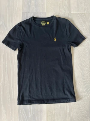 Svart t-shirt från Polo Ralph Lauren - Klassisk svart t-shirt från Polo Ralph Lauren med gul broderad logga på bröstet. Modellen är slim fit och har rund halsringning samt korta ärmar. Tillverkad i mjuk bomull som känns skön mot huden. Perfekt basplagg med stilren design. Vid intresse är det bara att höra av sig. Pris går att diskutera 