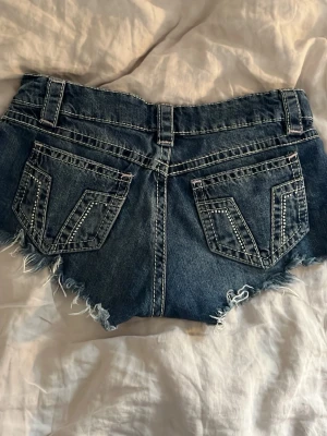 Blå jeansshorts med slitningar - Säljer ett par blå jeansshorts med råa, fransiga kanter och slitna detaljer framtill. Shortsen har bakfickor med dekorativa sömmar och bälteshällor. Perfekta för sommaren. Är storlek XXS i 14+ men känns som en XS-S