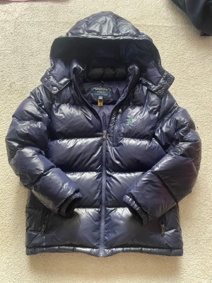 Mörkblå pufferjacka från Polo Ralph Lauren - Snygg mörkblå pufferjacka från Polo Ralph Lauren med glansig finish och broderad grön logga på bröstet. Jackan har huva, dragkedja framtill och två sidofickor med dragkedja. Perfekt för kalla vinterdagar och riktigt trendig look.