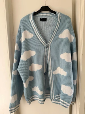 Blå kofta med molnmönster ASOS DESIGN - Superfin blå kofta från ASOS DESIGN med vita moln över hela plagget. Koftan är ribbstickad, har v-ringning och stängs med två knappar framtill. Mjuk och oversized.