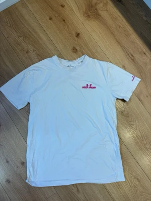 Under Armour t-shirt - Tröjan har en liten fläck på bild 2 har jag jag markerat fläcken.                                     Vit t-shirt från Under Armour med korta ärmar och rosa logga på bröstet samt ärmen. Klassisk passform och rund halsringning. Perfekt för dig som gillar sportig och enkel stil.