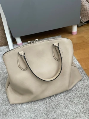 Beige handväska med dubbla handtag i äkta läder.(kom med prisförslag!!) - Stilren beige handväska i mjukt material med rundad form och dubbla handtag. Väskan har dragkedja upptill och minimalistisk design utan synliga loggor. Perfekt för dig som gillar enkel och klassisk stil.
