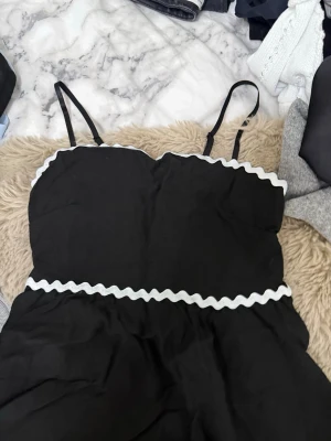 Svart topp med vita detaljer - Snygg svart topp med smala axelband och vågiga vita band längs byst och midja. Klänningen har en enkel och stilren design som passar perfekt till sommaren. Materialet känns lätt och bekvämt.