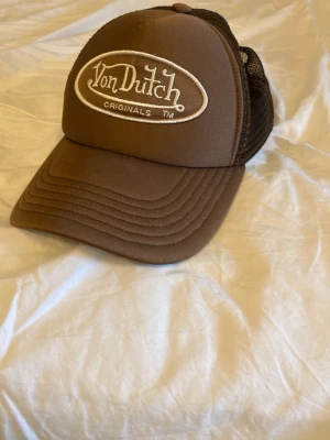 Brun Von Dutch keps - Säljer en brun truckerkeps från Von Dutch med vit broderad logga framtill. Kepsen har klassisk böjd skärm och mesh baktill för extra ventilation. Justerbar passform med snapback. Perfekt för dig som gillar retrostil och streetwear. Priset är diskuterbart skriv om ni har några frågor.