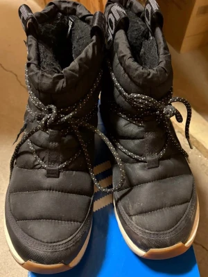 Svarta boots - Säljer ett par svarta boots med quiltad design och tjocka snören. Skorna har en vadderad ovandel och mjukt foder för extra värme. Sulan är vit med svarta detaljer och loggan syns tydligt på skaftet. Perfekta för kalla vinterdagar. OBS: små i storleken