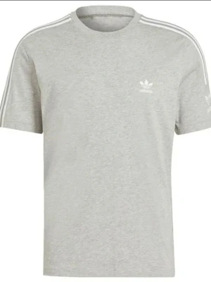 Grå Adidas t-shirt med vita ränder - Snygg grå t-shirt från Adidas med klassiska vita ränder längs axlarna och det ikoniska trefoil-loggan på bröstet. T-shirten är i mjuk bomull och har en normal passform med korta ärmar. Perfekt för en sportig och avslappnad stil.