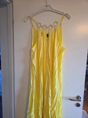 Gul klänning, nyskick, stl XL - Lång gul klänning med tunna axelband och dekorativa ringar framtill. Klänningen har en lös passform och är tillverkad i ett lätt tyg, perfekt för varma dagar. Volangdetalj längst ner ger extra rörelse och sommarkänsla.