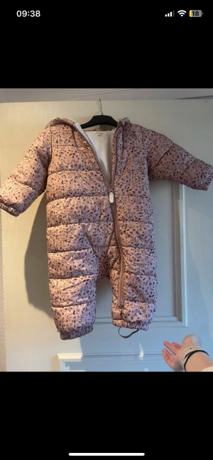 Rosa blommig dunoverall med huva - Söt rosa dunoverall med små blommor, hel dragkedja och huva. Overallen har resår vid ärm- och benslut samt mjukt innerfoder. Perfekt för kalla vinterdagar och håller värmen riktigt bra. Fatts en gummiband vid foten 