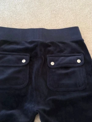 Mörkblå velourbyxor Juicy Couture - Mörkblå mjukisbyxor i velour från Juicy Couture med två bakfickor med lock och silverfärgade knappar. Byxorna har broderad JC-logga på sidan och bred resår i midjan. Perfekta för en chill och trendig look. Betalning via vanlig swish! 
