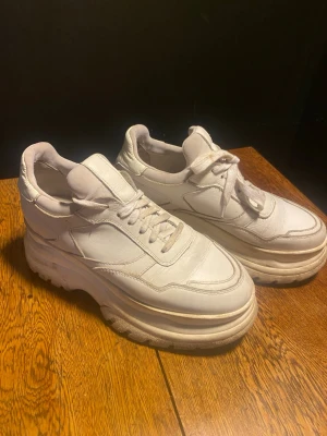 Chunky vita sneakers med platåsula - Säljer ett par chunky vita sneakers med kraftig platåsula och klassisk snörning. Skorna har en modern och sportig look med detaljerade sömmar och rundad tå. Tillverkade i vitt läderliknande material som ger en clean och trendig vibe.