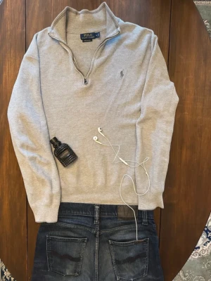 Grå halvzip tröja från Polo Ralph Lauren - Snygg grå tröja från Polo Ralph Lauren med halv dragkedja och klassisk broderad logga på bröstet. Tröjan har hög krage och är tillverkad i mjukt bomullsmaterial. Perfekt till jeans eller chinos för en clean look.