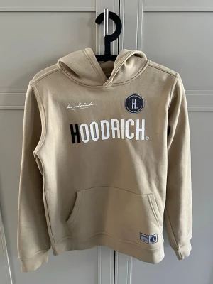 Beige hoodie från Hoodrich - Snygg beige hoodie från Hoodrich med stor logga framtill och tryck på ryggen. Hoodien har huva, känguruficka och ribbade muddar. Tillverkad i mjukt material som känns skönt mot huden. Perfekt för en avslappnad streetwear-look.