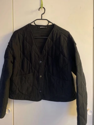 Svart quiltad jacka från Gina Tricot - Stilren svart quiltad cardiganjacka från Gina Tricot med tryckknappar framtill och två stora fickor. Jackan har V-ringning, lång ärm och ett vågigt quiltat mönster som ger en trendig look. Perfekt att slänga över en hoodie eller t-shirt.