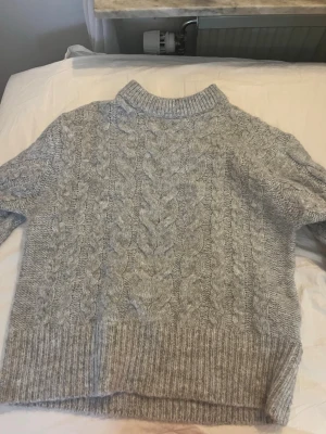 Grå kabelstickad tröja med polokrage - Mysig grå kabelstickad tröja med hög ribbad polokrage och breda muddar. Tröjan har ett klassiskt flätmönster framtill och är perfekt för kyliga dagar. Passar dig som gillar en avslappnad men ändå stilren look.