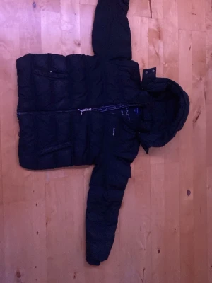 Svart pufferjacka från GANT - Säljer en svart pufferjacka från GANT med huva och dragkedja framtill. Jackan har en quiltad design och två sidofickor med dragkedja. Perfekt för kalla vinterdagar och riktigt skön att bära.