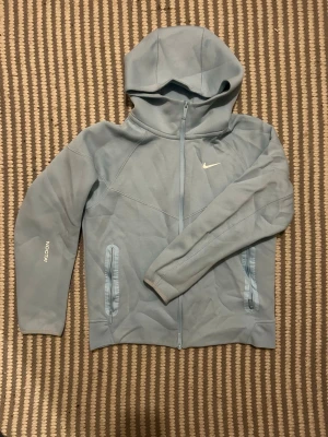 Ljusblå Nike NOCTA hoodie - Snygg ljusblå hoodie från Nike NOCTA-kollektionen med dragkedja framtill, två fickor och huva. Diskret Nike-logga på bröstet och NOCTA-tryck på ärmen. Tillverkad i mjukt material som känns skönt mot huden. Perfekt för en avslappnad och sportig stil.