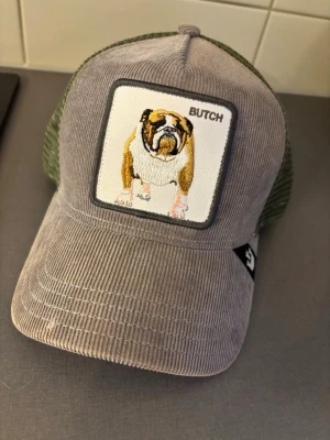 Grå keps med bulldogpatch Goorin Bros - Säljer en grå keps från Goorin Bros med en broderad bulldog och texten 'BUTCH' på framsidan. Kepsen har mesh i grönt baktill och är tillverkad i manchestertyg framtill. Justerbar passform och cool patch framtill gör den unik.